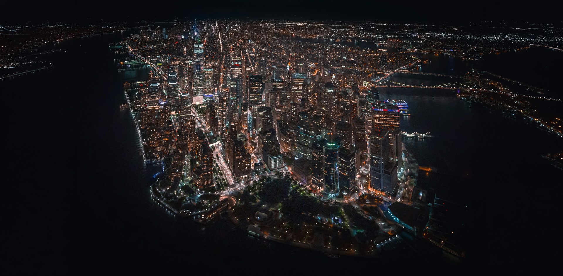 Aerial night view of Manhattan looking south from above the city, the financial district illuminated against the dark water of the surrounding harbor.