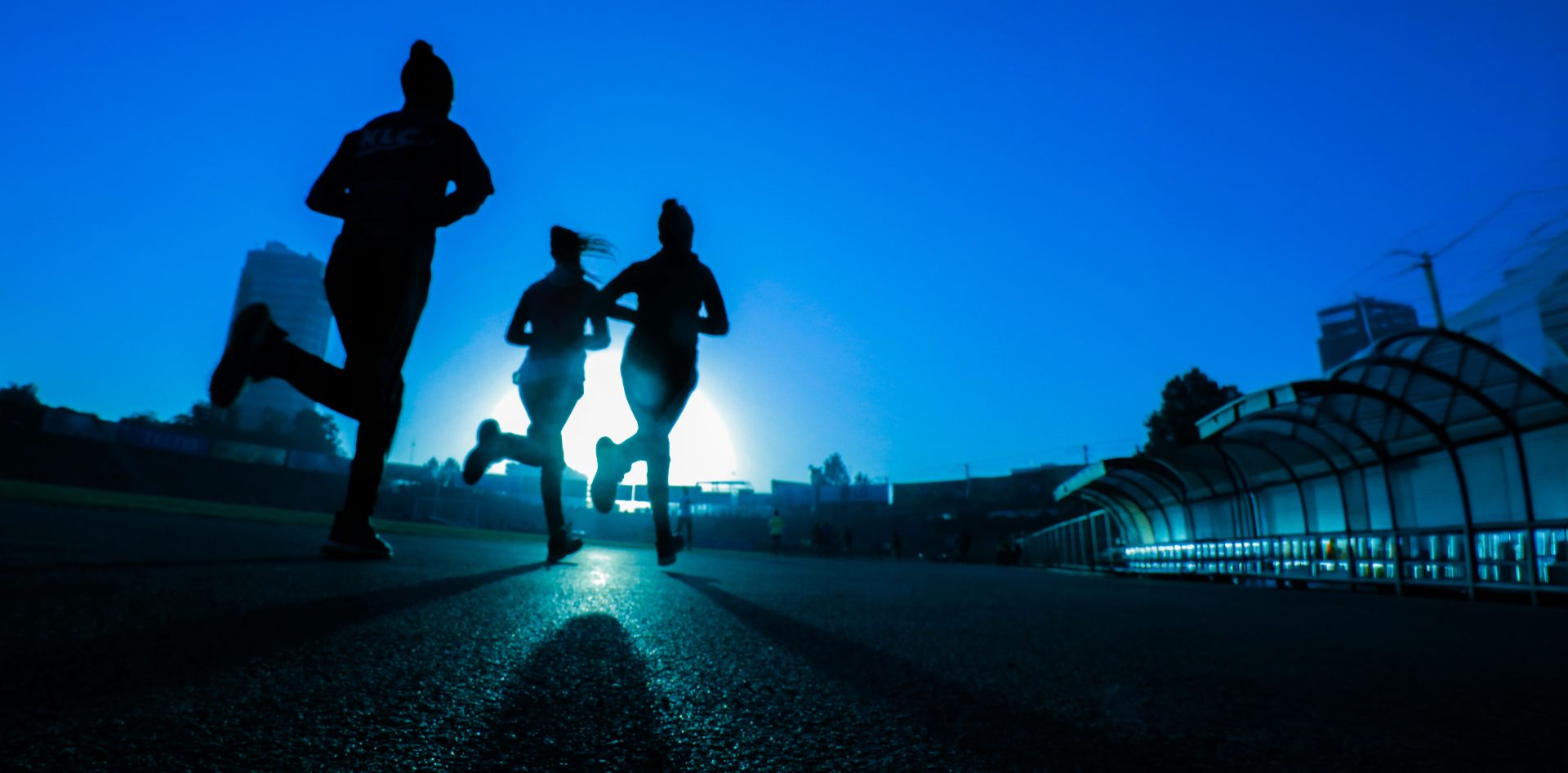 Three runners silhouetted against a vivid blue twilight sky, mid-stride on a track at sunrise.
